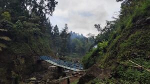 Jembatan besi biru di tengah hutan lebat dan lembah berbatu di kawasan Tirta Sena - Curug Muntu, dikelilingi pepohonan tinggi dan tebing hijau.