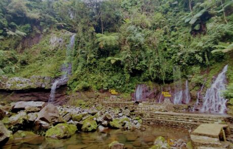 Pemandangan Curug Muntu dan Tirta Sena di Desa Ketenger, dengan air terjun kecil yang mengalir dari tebing hijau lebat, dikelilingi batu alam dan jalur tangga wisata
