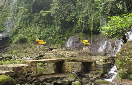 Panorama dua air terjun, Curug Muntu dan Tirta Sena, yang mengalir berdampingan di tengah hutan tropis lebat di Desa Ketenger, dengan papan nama kuning yang mencolok di antara rimbunan hijau